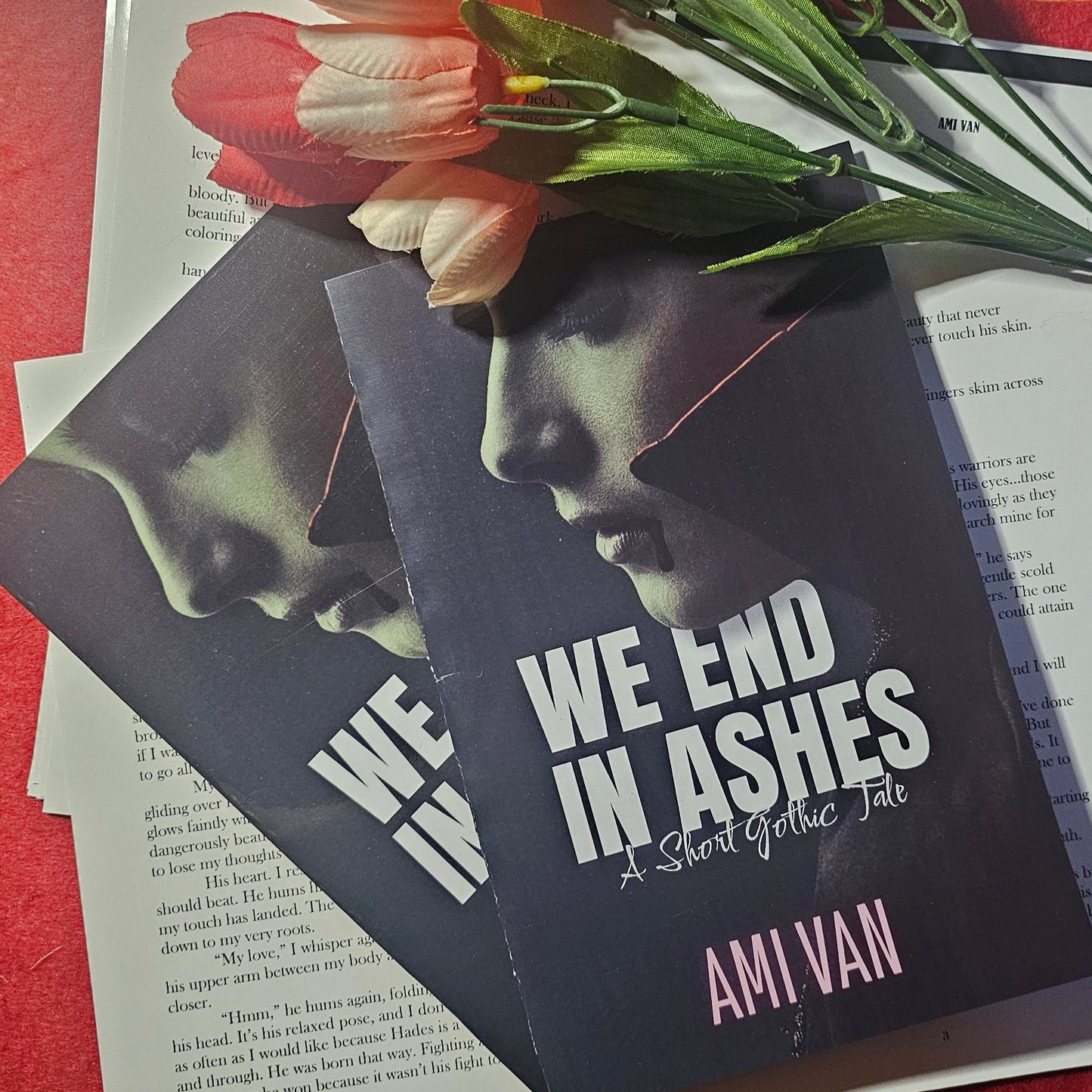 Book titled 'We End in Ashes' by Ami Van on a red surface with flowers and stationery items.