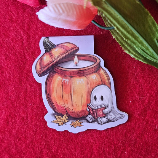 A magnetic bookmark with an illustration of a cute ghost sitting in front of a large pumpkin while reading a book. 