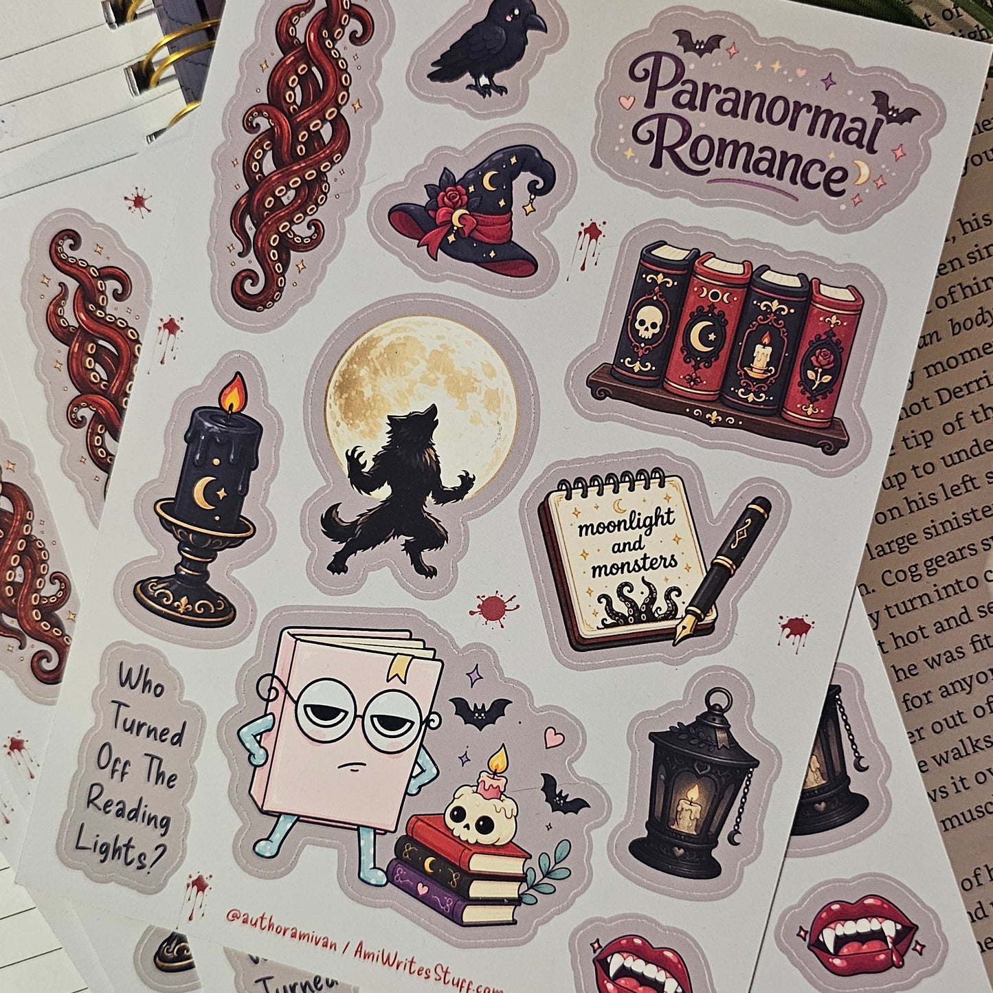 A sheet of decorative stickers featuring gothic and horror-themed illustrations, including a crouching werewolf under a full moon, a witch hat, books, a candle, and the words 'Paranormal Romance,' placed on top of an open notebook.