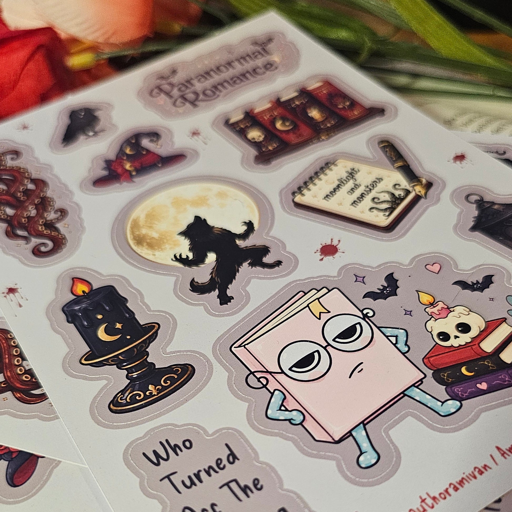 A close-up of sticker sheets featuring Halloween-themed illustrations, including a black cat silhouette, a candle holder with a crescent moon, a book character with glasses, and spooky text, arranged on a white surface with decorative flowers in the background.
