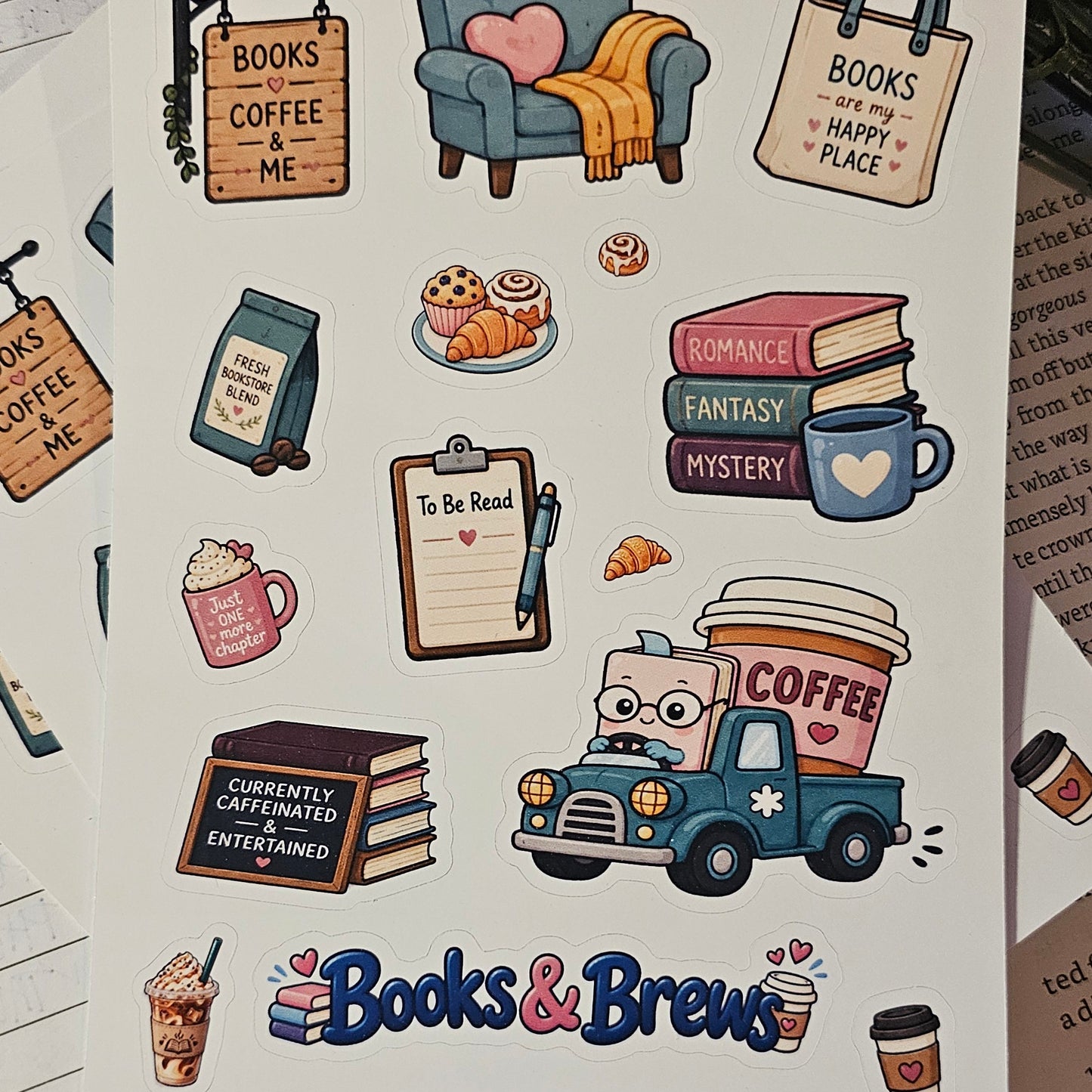 A sheet of stickers featuring cozy books and coffee-themed illustrations, including a blue armchair with a yellow blanket, a coffee truck, books, mugs, and signs reading 'Books Coffee Me' and 'Books Happy Place.' The sheet is placed on top of other notebooks and papers on a red fabric surface with flowers in the background.