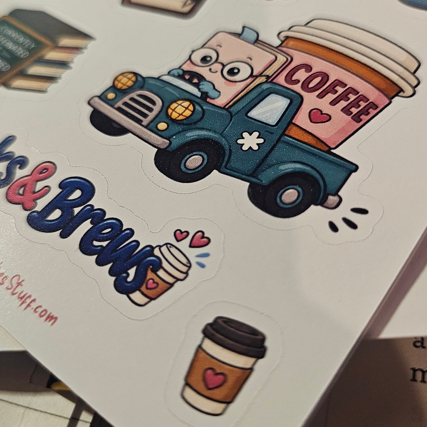 A close-up of a sticker sheet featuring a cartoon coffee truck with the word 'COFFEE' on its side, along with coffee cup stickers, books, and decorative text stickers, photographed on a white sheet held by a gold binder clip.
