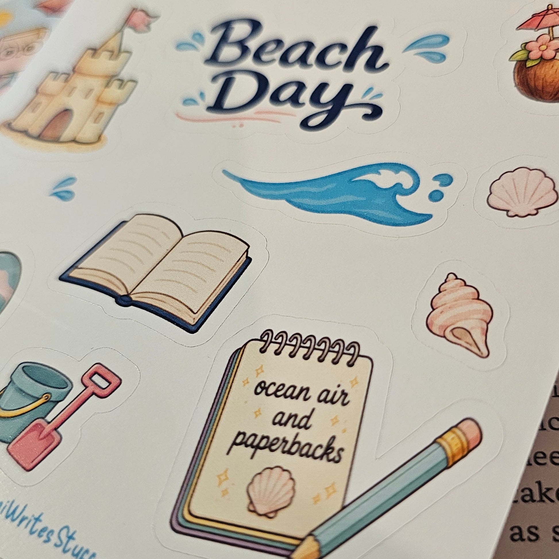 A close-up of a sticker sheet featuring beach-themed illustrations, including a sandcastle, beach bag, stack of books, umbrella, waves, seashells, a pencil, and a notebook that reads 'ocean air and paperbacks.' The sheet is photographed on top of other printed materials.