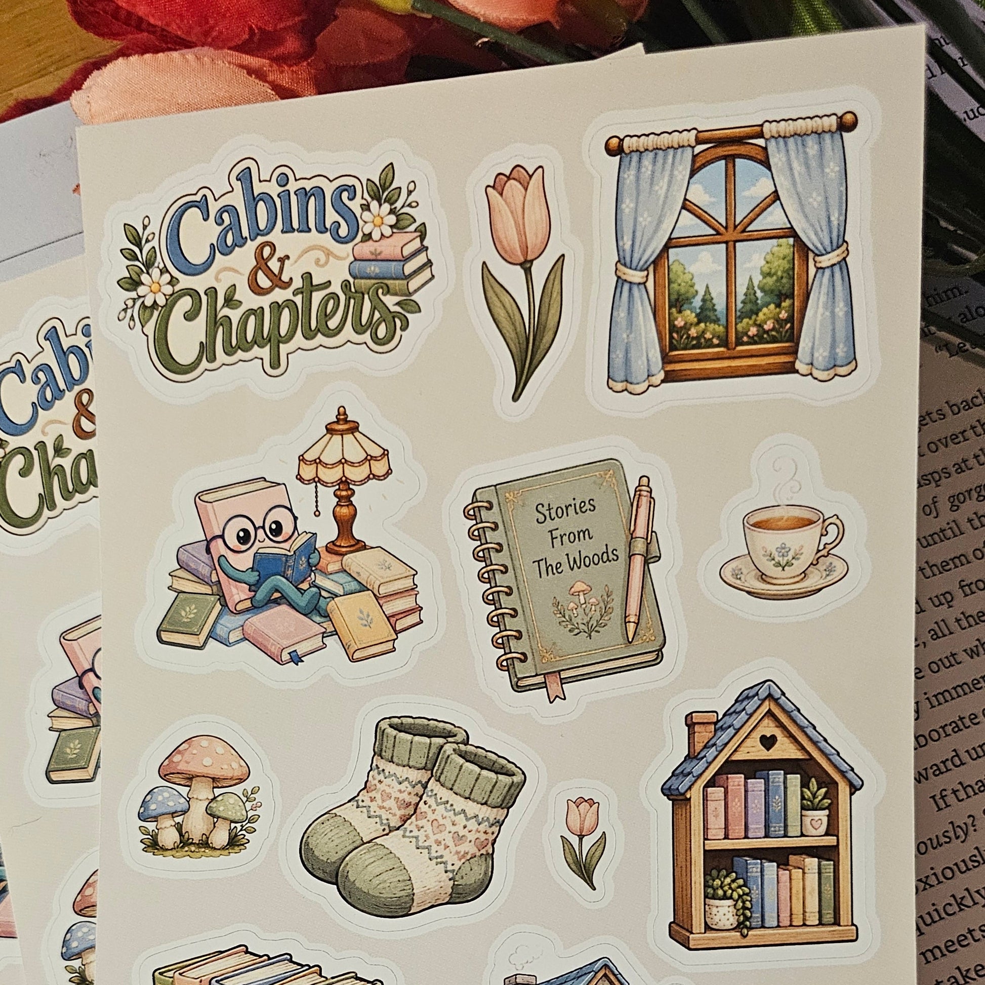 A sheet of illustrated planner stickers with cozy cabin-themed designs, including a window, books, socks, a small house, a cart, flowers, and text reading 'Cabins & Chapters,' photographed on top of lined notebook pages.