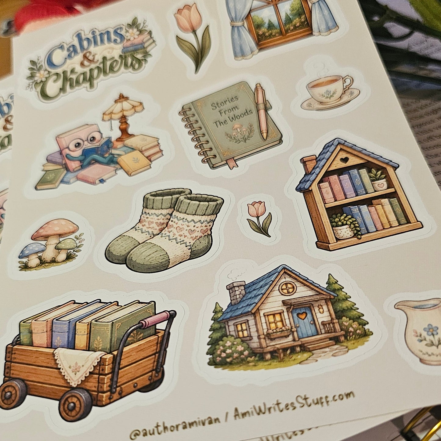 A sheet of illustrated stickers featuring cozy cabin-themed designs such as a small house, books in a cart, knit socks, a window, a teacup, and a notebook, photographed on top of other sticker sheets with flowers in the background.