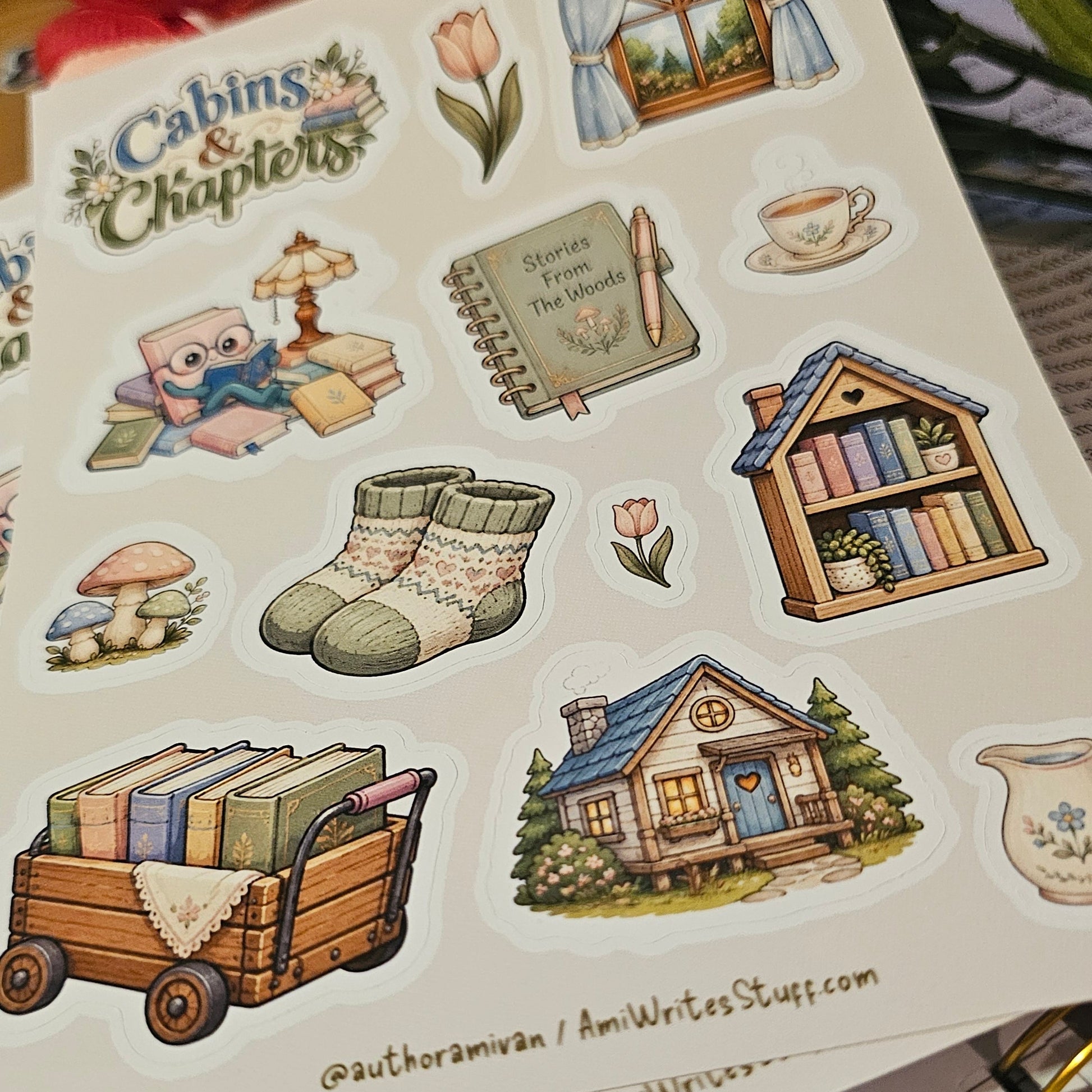 A sheet of illustrated stickers featuring cozy cabin-themed designs such as a small house, books in a cart, knit socks, a window, a teacup, and a notebook, photographed on top of other sticker sheets with flowers in the background.