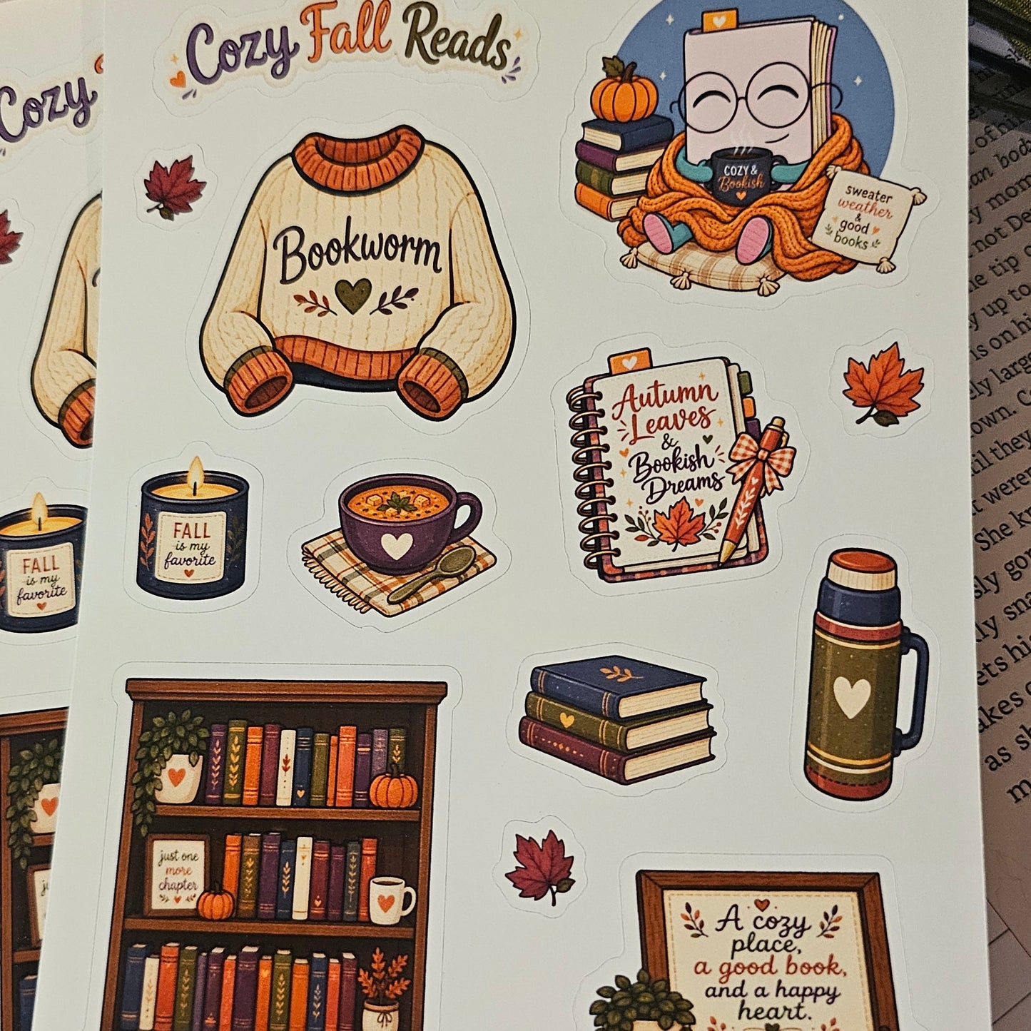 A sheet of illustrated stickers featuring cozy autumn and book-themed designs, including a sweater that says 'Bookworm,' a reading character, books, a bookshelf, candles, and fall leaves, photographed on top of other sticker sheets with flowers and a book in the background.