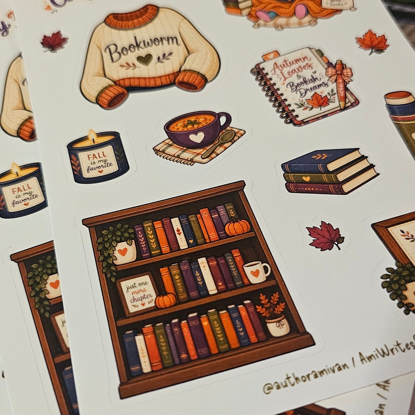 A stack of printed sticker sheets featuring cozy fall-themed illustrations, including a sweater that says 'Bookworm', books, a bookshelf, autumn leaves, and handwritten-style titles like 'Cozy Fall Reads'.