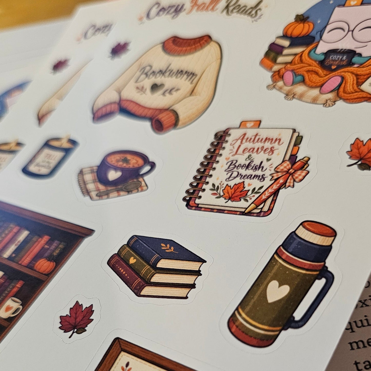 A close-up of a sticker sheet featuring cozy autumn-themed illustrations, including a sweater that says 'Bookworm', a notebook with 'Autumn Leaves British Dreams', a mug, books, a thermos, and a sign reading 'A cozy place, a good book, and a happy heart'.