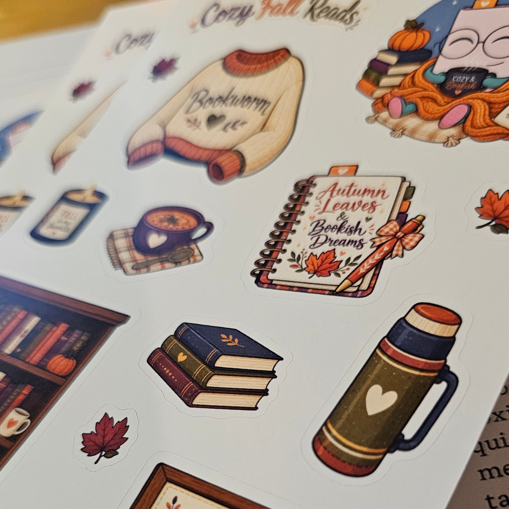 A close-up of a sticker sheet featuring cozy autumn-themed illustrations, including a sweater that says 'Bookworm', a notebook with 'Autumn Leaves British Dreams', a mug, books, a thermos, and a sign reading 'A cozy place, a good book, and a happy heart'.