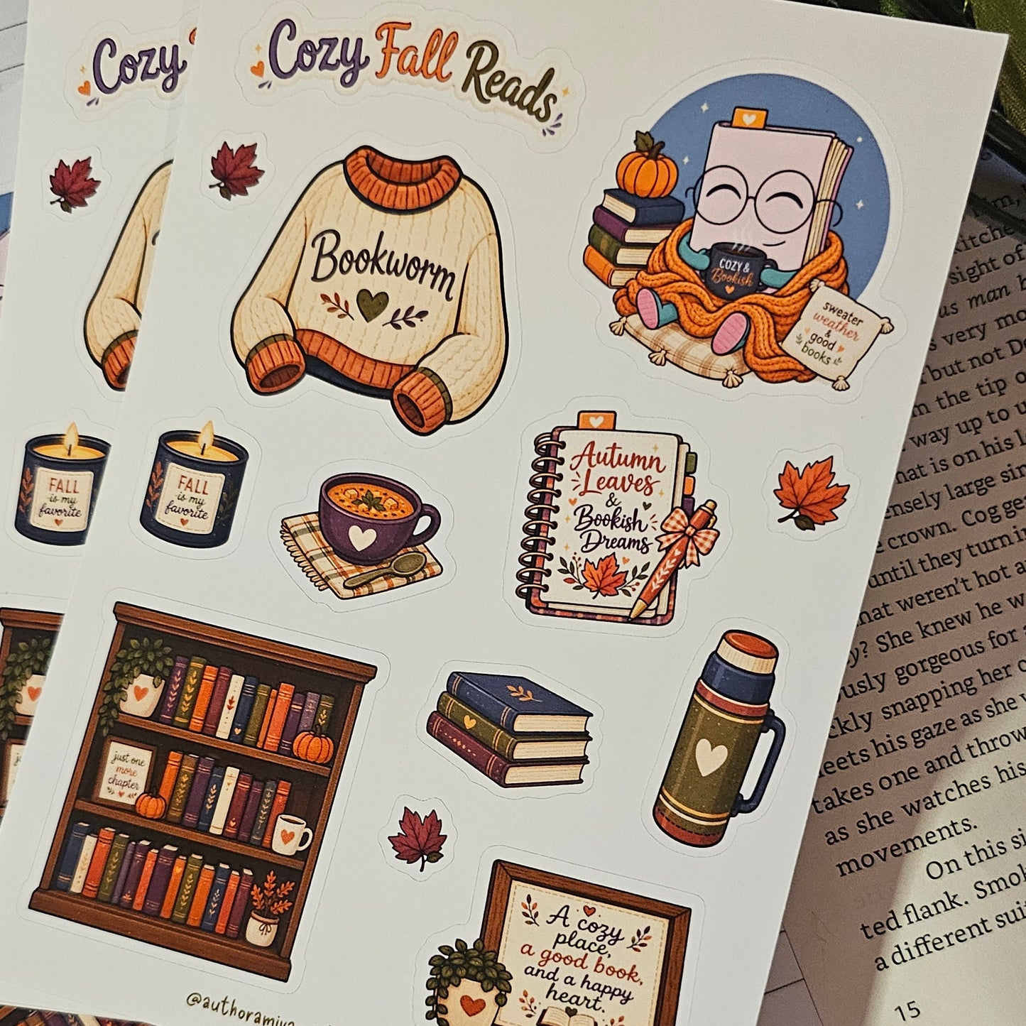 A sheet of cozy autumn-themed bookish stickers displayed on top of other sticker sheets and an open book, resting on a red fabric surface with a wooden table edge visible.
