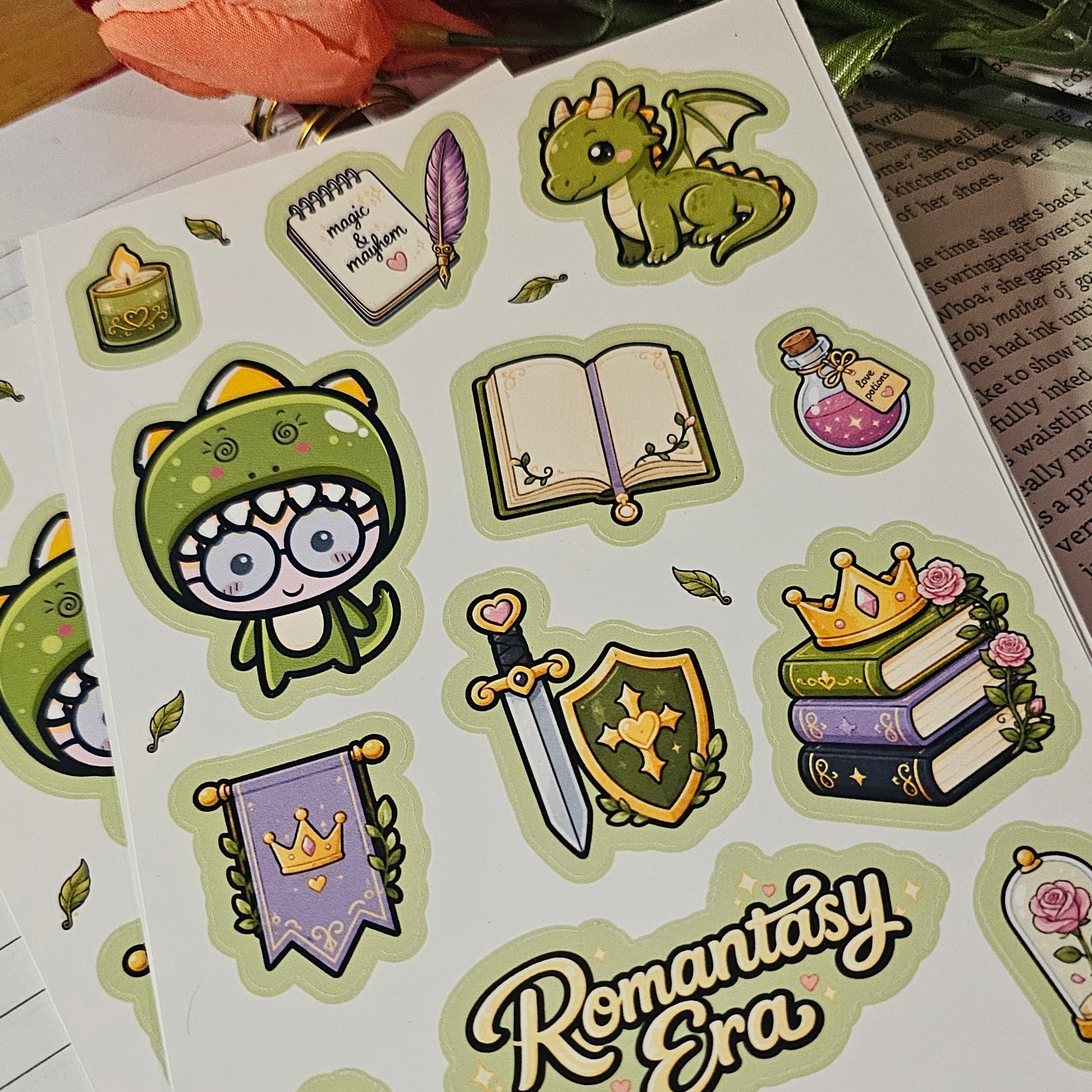A sheet of cute fantasy-themed stickers displayed on an open lined notebook, with a blurred pink flower in the background. The stickers feature a green monster, books, a sword, a potion bottle, a crown, and the text 'Romantasy Era'.