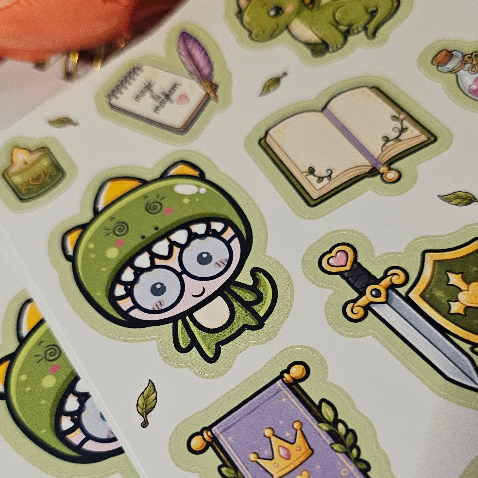 A close-up of sticker sheets featuring cute green dragon illustrations and themed stickers, including a dragon in a knight helmet, an open book, a sword, a crown banner, and small leaves, arranged on a white surface with a lined paper and blurred flowers in the background.