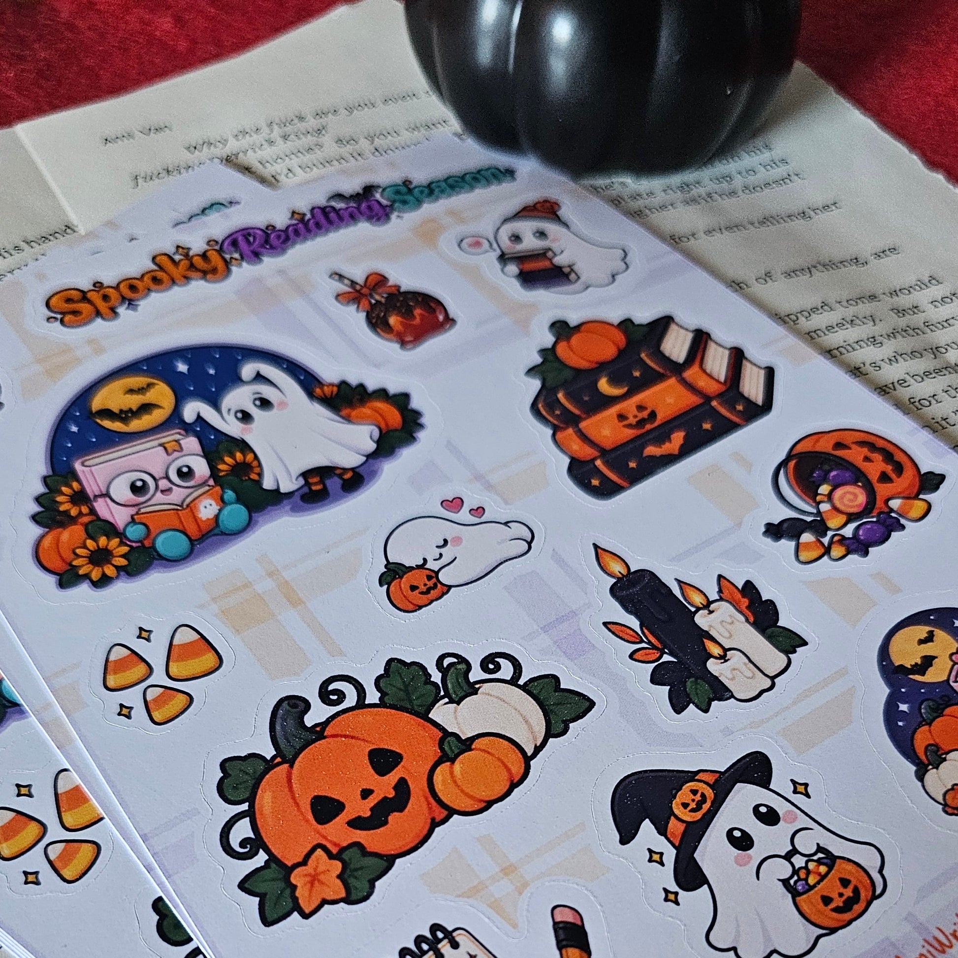 A sheet of cute Halloween-themed stickers laid on top of open book pages, with more sticker sheets and a black decorative pumpkin visible in the background.