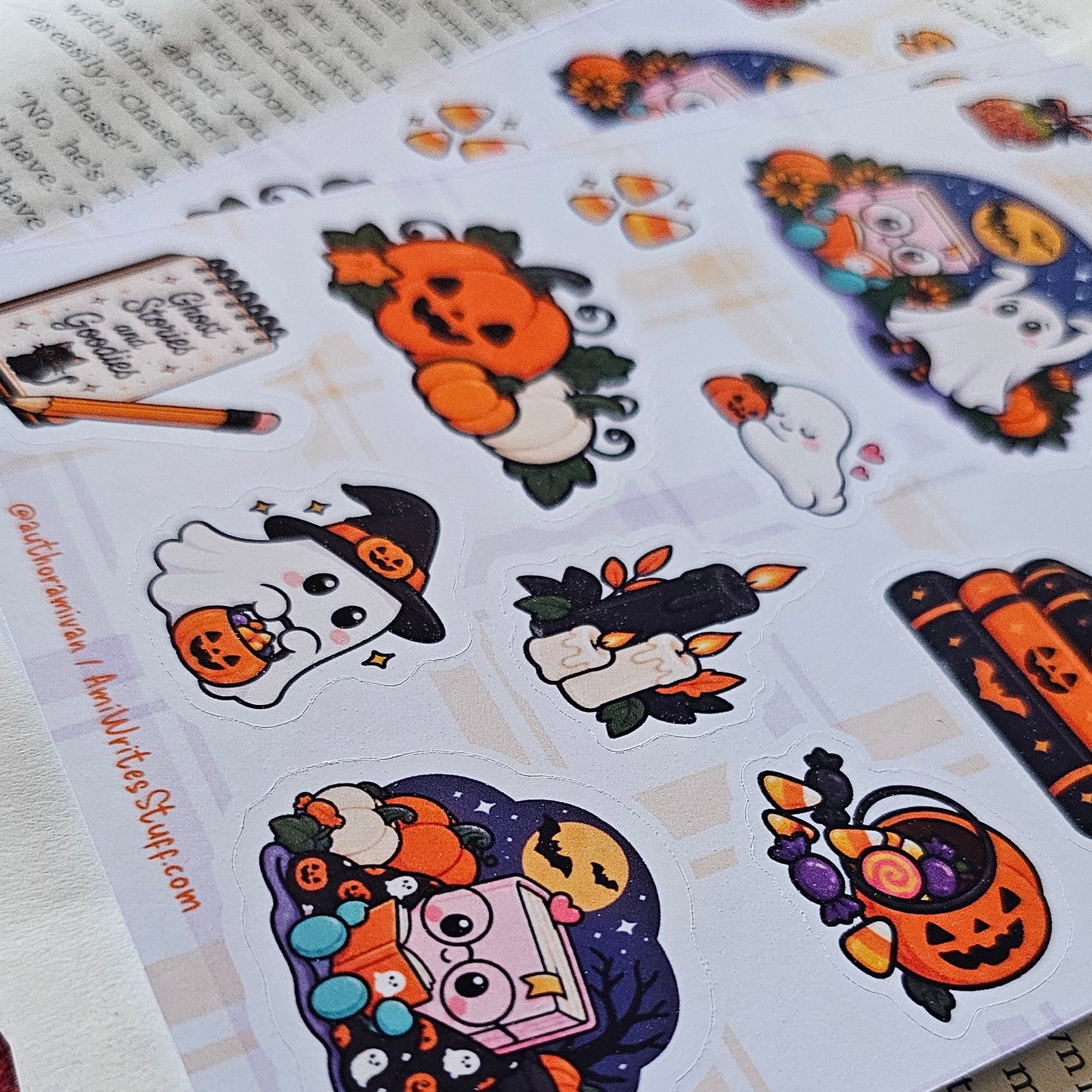 A printed sheet of Halloween-themed stickers laid on top of an open book. The stickers feature cute cartoon illustrations including pumpkins, ghosts, a witch, a cat, and candy in orange, purple, white, and black.